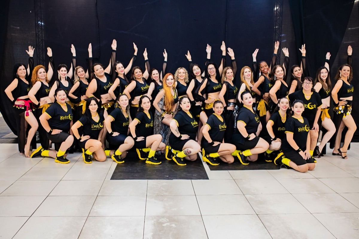 Turma do EvoLua em pose de grupo com braços erguidos, uniforme preto e amarelo, telão preto ao fundo