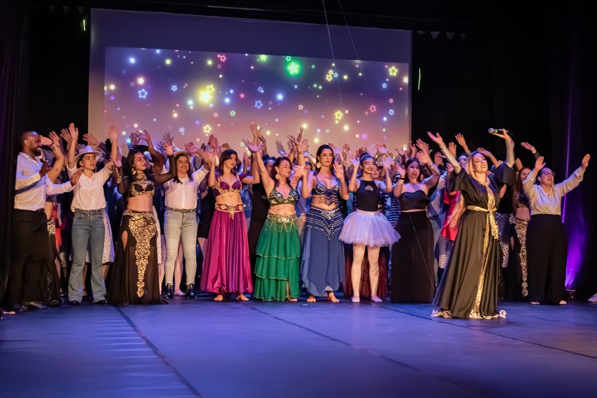 Turma do EvoLua comemorando no palco após apresentação, alunas de braços erguidos aplaudindo, telão com efeito de estrelas ao fundo
