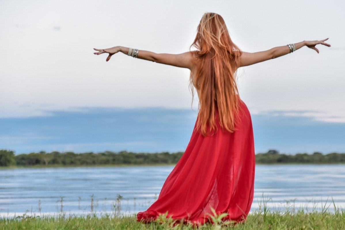 Viviane Lua de costas, vestido vermelho longo, braços abertos, à beira de um lago