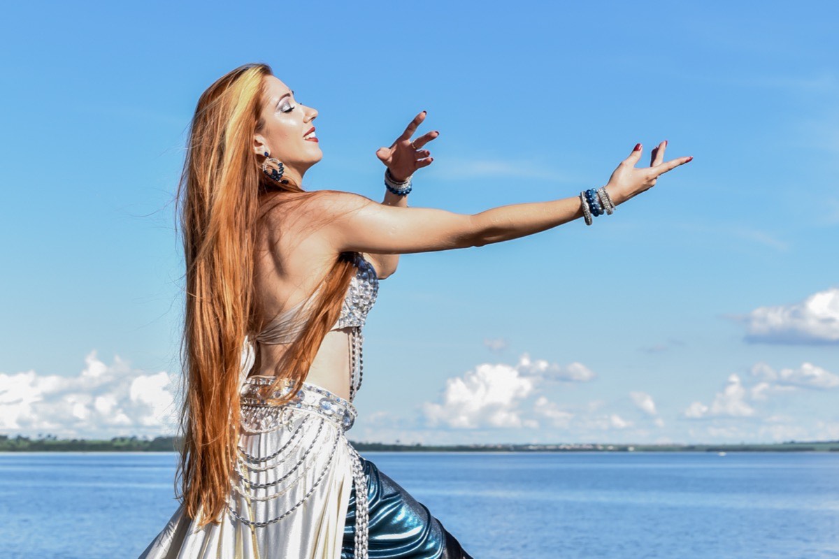 Viviane Lua de perfil dançando à beira-mar, figurino prateado e azul de dança do ventre, céu azul