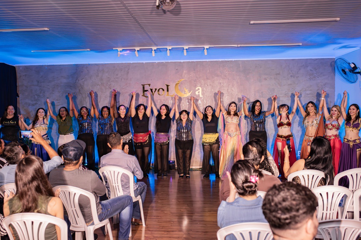 Grupo de alunas em fila no palco, mãos dadas erguidas, backdrop EvoLua, plateia sentada