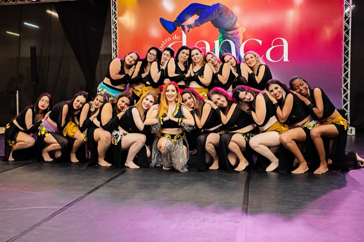 Grupo de alunas do EvoLua posando agachadas no palco, figurino de dança do ventre preto e dourado, backdrop Mostra de Dança